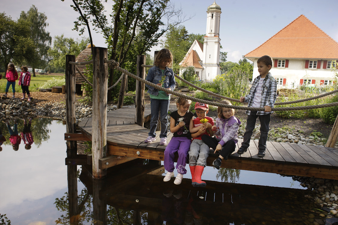 Kinder sitzen auf einem Steg und lassen die Füße ins Wasser baumeln